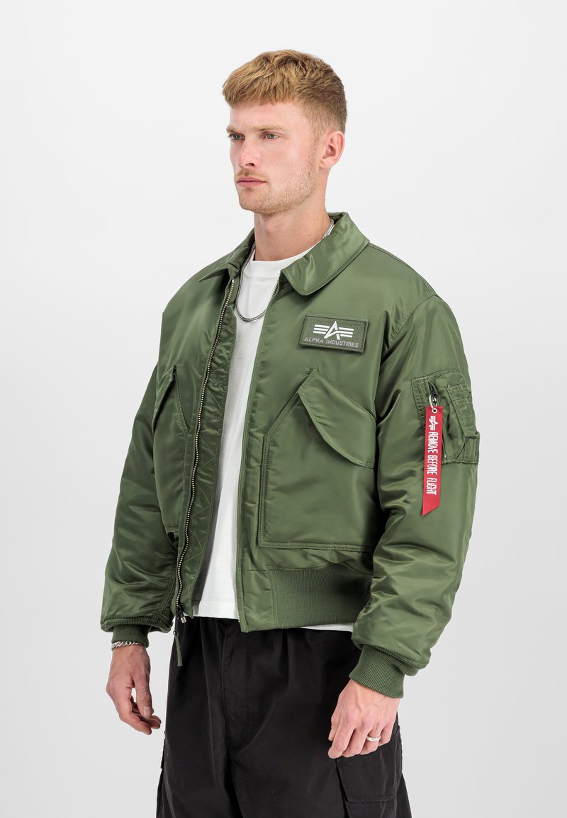 Mand iført grøn Alpha Industries bomberjakke med forlommer og rød "Remove Before Flight"-mærke på ærmet, hvid skjorte og sorte bukser.