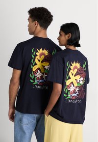 Marineblå bomulds t-shirts har farverige blomstermotiver, herunder en solsikke og en gul blomst, med brandnavnet "L’Antidote" trykt nedenfor.