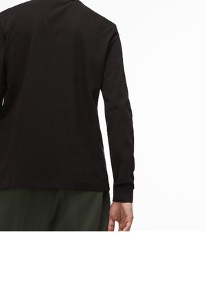 Vue de dos d'une personne portant une chemise noire à manches longues et un pantalon vert foncé, debout avec un bras détendu le long du corps.