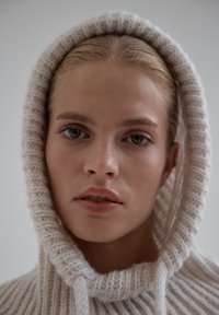 Jasny, dzianinowy hoodie w kolorze kremowym z fakturą prążkowaną, z kapturem na sznurki i głębokim kapturem. Model ma neutralny wyraz twarzy.