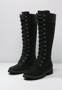 Svarta knähöga stövlar i mocka, med kontrasterande svarta snören och hexagonala snörhål, samt en robust gummisul och texturerad yta.