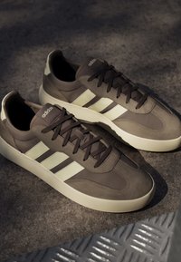 Een paar bruine Adidas sneakers met witte strepen en zolen op een gestructureerd grijs oppervlak bij zacht licht.
