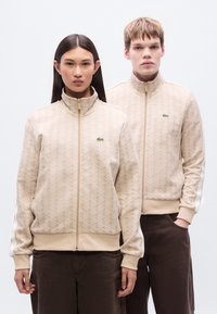 Μπεζ μπουφάν με φερμουάρ και γεωμετρικό σχέδιο, με ψηλό γιακά και πράσινο λογότυπο στο αριστερό στήθος. Διαθέτει ριμπ μανσέτες και τελείωμα.