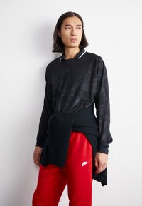 Černá košile s dlouhými rukávy a jemným vzorem, bílý límeček a červené tepláky s bílým logem Nike, doplněné tmavým svetrem uvázaným kolem pasu.