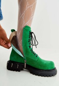 Groene suède enkellaars met een zwarte chunky zool, vetersluiting aan de voorkant, ritssluiting aan de zijkant en een contrasterende lichtbruine hak. Visnetkousen zichtbaar.