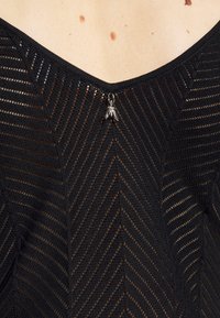 Czarny dzianinowy top z wzorem w zygzak, z przezroczystymi sekcjami, dekoltem w kształcie litery V oraz metalicznym akcentem w centrum.