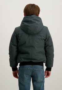 Donkergroene gewatteerde jas met capuchon, elastische manchetten en een zwarte tailleband, gecombineerd met lichtblauwe spijkerbroek.