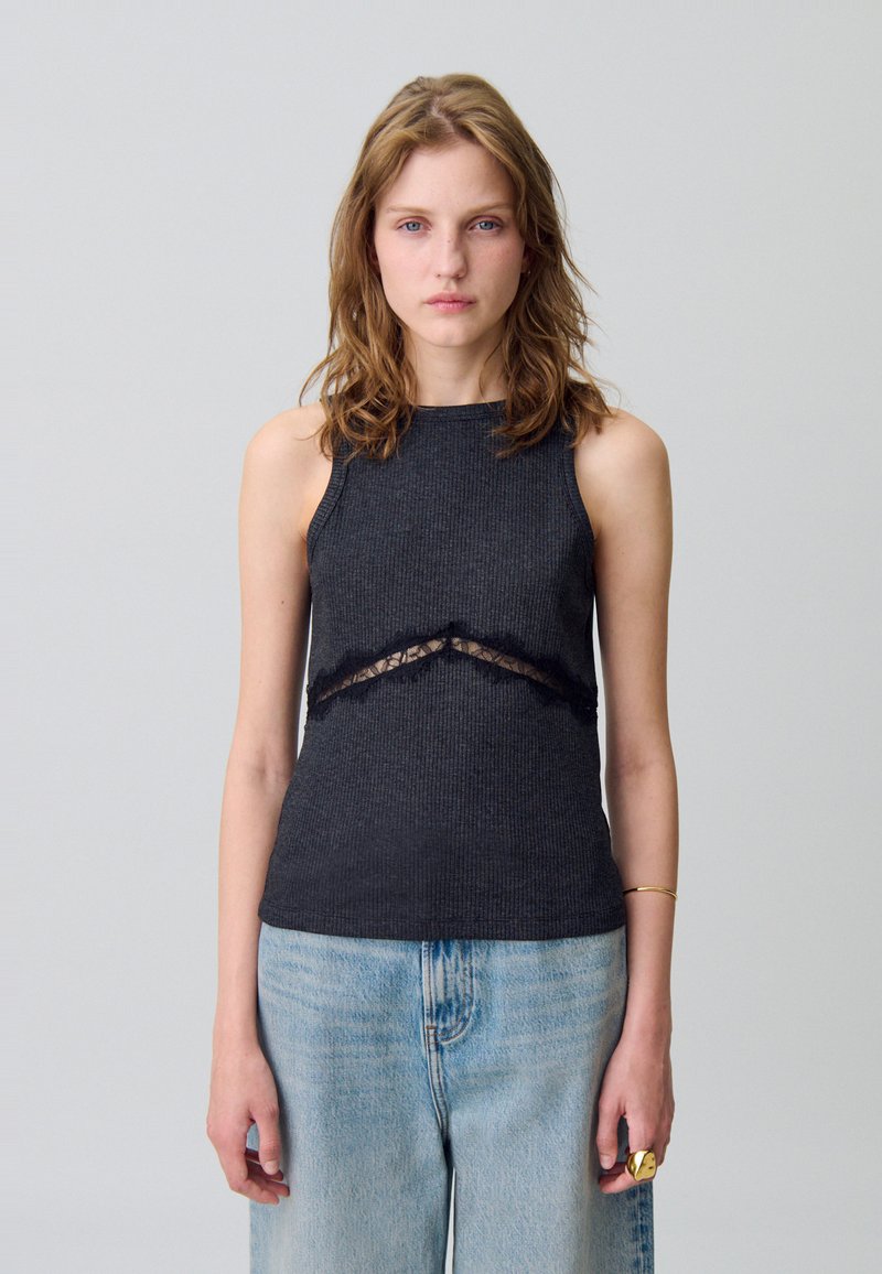 La camiseta de tirantes negra con canalé presenta un detalle de encaje recortado debajo del busto. Se combina con jeans de mezclilla azul claro, creando un look casual.