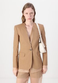 Bruine linnen blazer met één knoop, gelapte revers, en twee voorkockets; gecombineerd met een transparante lichtbruine top en shorts.