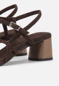 Καφέ σουέντ σανδάλια με χοντρό τακούνι, λουράκια που διασταυρώνονται και ασημένια αγκράφα σε ανοιχτό φόντο.
