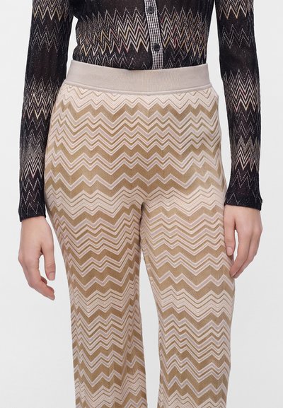 Personne portant un pantalon beige et rose clair à motif zigzag et un haut noir transparent à manches longues avec un motif zigzag multicolore et des boutons.