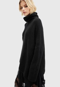 Melns turtlenecks džemperis, kas izgatavots no maiga, strukturēta materiāla, ar brīvu piegriezumu un ribotu apakšmalu, apvienots ar caurspīdīgu melnu mežģīņu svārku.