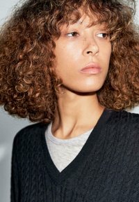 Maglione nero lavorato a maglia con scollatura a V profonda, indossato sopra una maglietta grigia. Motivo testurizzato visibile, con capelli castani ricci di lunghezza media.