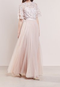 Robe maxi avec un haut transparent orné, featuring des manches volantes, une taille cintrée et une jupe en tulle rose pâle fluide. Chaussures scintillantes visibles.