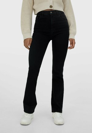 Personne portant un jean noir taille haute, des baskets blanches et un cardigan court en maille beige avec un bouton à l'avant.