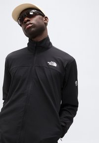 Άνδρας με μπεζ καπέλο, μαύρα γυαλιά ηλίου και μαύρο τζάκετ με φερμουάρ και λογότυπο The North Face, πόζαρε μπροστά σε απλό ανοιχτό φόντο.