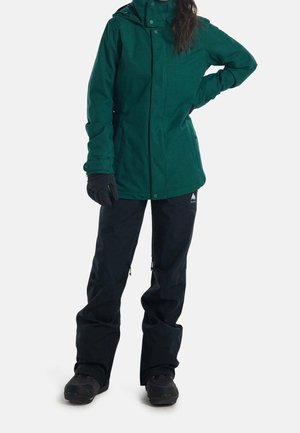 Veste verte, texturée, avec capuche et fermeture à pression, associée à un pantalon imperméable noir avec des zips aux chevilles et des chaussures noires.