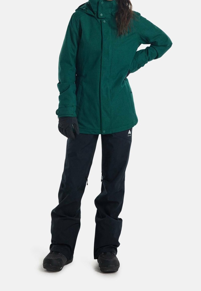 Grüne, strukturierte Jacke mit Kapuze und Druckknopfverschluss, kombiniert mit schwarzen wasserdichten Hosen mit Reißverschlüssen an den Knöcheln und schwarzen Schuhen.