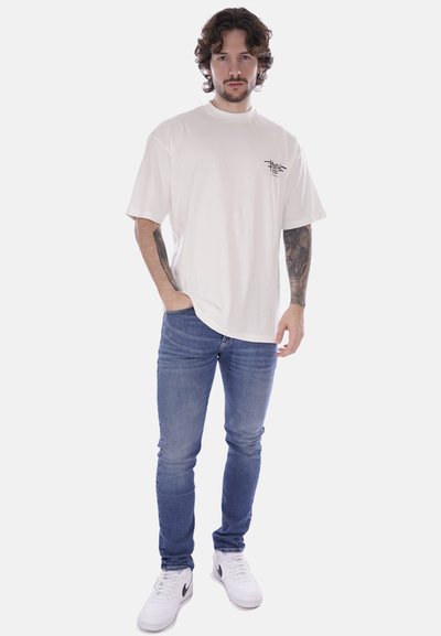 T-shirt bianca in cotone con maniche corte e piccola grafica nera sul lato sinistro del petto, abbinata a jeans di denim e sneakers bianche.
