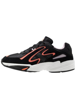 Black athletic shoe with a suede and mesh upper, pink accents, textured sole, and a chunky white midsole design. Features contrasting stripes.