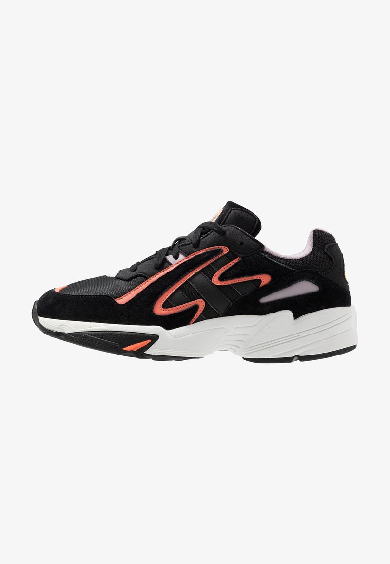 Black athletic shoe with a suede and mesh upper, pink accents, textured sole, and a chunky white midsole design. Features contrasting stripes.