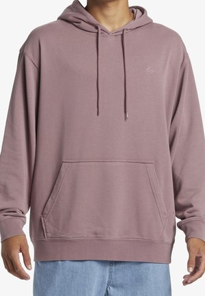 Osoba ubrana w mauve-bluzę z kapturem i kieszenią z przodu oraz jasnoniebieskie dżinsy, widoczna od szyi do bioder na jednolitym tle.
