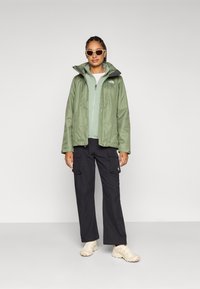 Jachetă impermeabilă verde cu fermoar, purtată peste un top verde deschis, asortată cu pantaloni cargo negri și adidași bej. Ochelarii de soare albi completează ținuta.