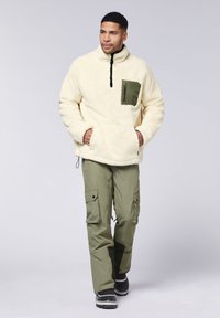 Cremfarbener Fleece-Pullover mit hohem Kragen, einer Fronttasche und olivgrünen Akzenten; kombiniert mit olivfarbenen Cargo-Hosen und schwarzen Stiefeln.