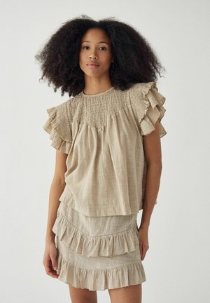 Žena s objemnými kučeravými vlasmi, ktorá má na sebe béžový vrúbkovaný top s krátkym rukávom a zodpovedajúcu vrstvenú vrúbkovanú sukňu, stojaca pred jednoduchým pozadím.