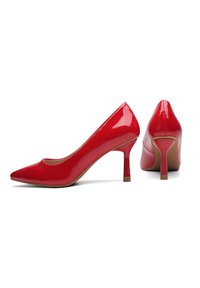 Tacchi alti in vernice rossa con punta affilata e sottile tacco a stiletto. Texture liscia e minimalismo nei dettagli, con interno beige.