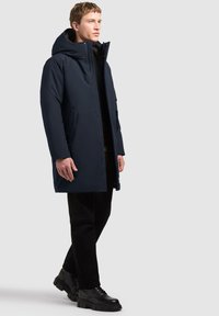 Homme portant un manteau d'hiver à capuche bleu marine, un pantalon noir et des chaussures noires épaisses marchant devant un fond uni.