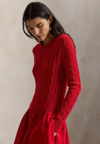 Γυναίκα με μακριά σκούρα μαλλιά που φοράει κόκκινο πλεκτό πουλόβερ με σχέδιο καλώδιο και ταιριαστή κόκκινη φούστα, κοιτάζοντας προς τα κάτω μπροστά σε απλό φόντο.