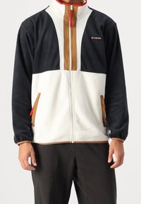 Giacca in pile con zip frontale, blocchi di colore in blu navy, crema e marrone, con accenti rossi e tasche laterali. Tessuto morbido, vestibilità rilassata.