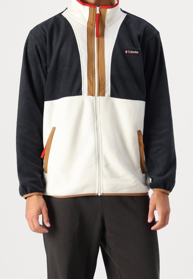 Giacca in pile con zip frontale, blocchi di colore in blu navy, crema e marrone, con accenti rossi e tasche laterali. Tessuto morbido, vestibilità rilassata.