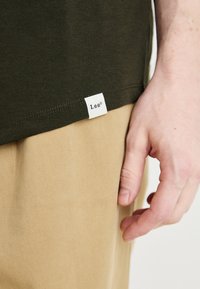Tricou din bumbac verde închis cu eticheta albă "Lee" pe tiv. Pantaloni bej cu o țesătură texturată. Prim-plan al unei mâini care atinge tricoul.