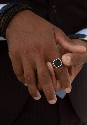 Zilveren vierkante ring met een zwarte center, gedragen aan een hand met een getextureerde, donkere huid, die een gevlochten zwarte armband toont.