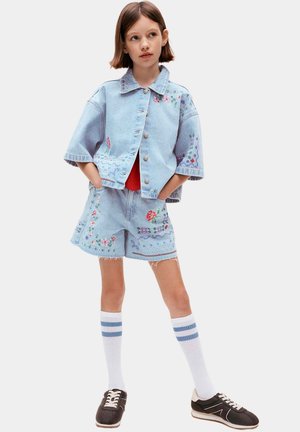 Junges Mädchen trägt eine hellblaue bestickte Jeansjacke und Shorts, weiße knielange Socken mit blauen Streifen und schwarze Turnschuhe und steht mit den Händen in den Taschen.