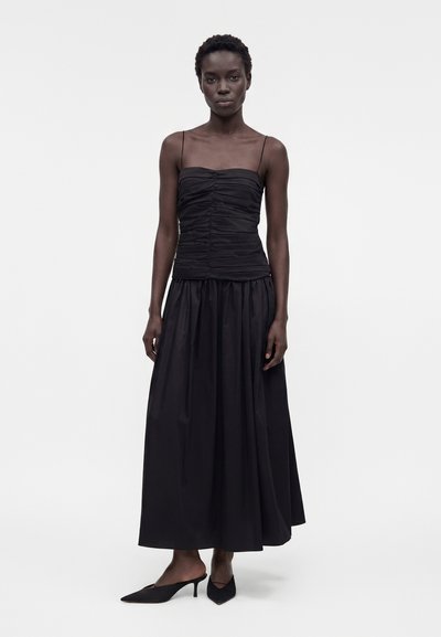 Γυναίκα που φορεί μαύρο τοπ με ραμμένες τιράντες και μακριά μαύρη φούστα που ρέει, με μαύρες μυτερές γόβες, στέκεται σε λευκό φόντο.