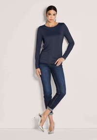 Vrouw in een donkerblauwe longsleeve, met donkerblauwe opgerolde jeans en zilveren hoge hakken, staande tegen een lichte achtergrond.