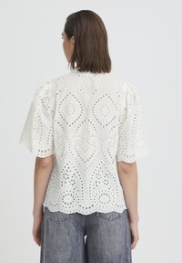 Femme aux cheveux bruns mi-longs portant un chemisier blanc à manches courtes en dentelle œillet et un jean denim gris, vue de dos.