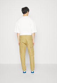 Khaki cargo kalhoty s bočními kapsami, kombinované s volným bílým tričkem, pohled zezadu. Minimalistický design, ležérní styl.