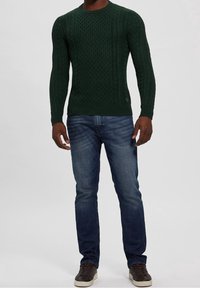 Green knitted sweater with textured cable pattern, crew neckline, paired with blue jeans and dark sneakers. No branding visible.