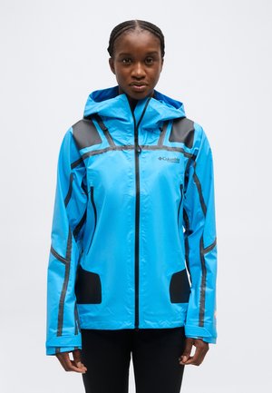 Femeie purtând o geacă impermeabilă Columbia Titanium albastră, cu glugă, accente negre și buzunare cu fermoar, stând în fața unui fundal deschis, simplu.