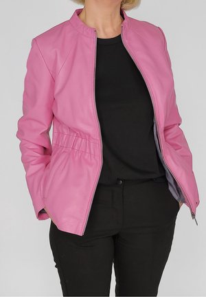 Femme portant une veste zippée rose par-dessus une chemise noire et un pantalon noir, les mains dans les poches de la veste, sur un fond gris.