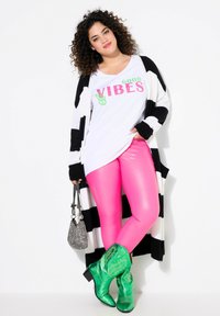T-shirt bianca con "GOOD VIBES" in rosa e verde, cardigan a righe bianche e nere, leggings rosa lucidi, stivali da cowboy verdi, borsa argentata.