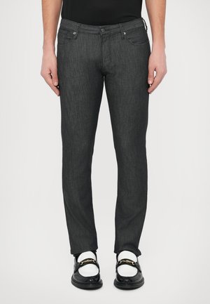 Homme portant un jean slim gris foncé, des chaussettes blanches et des mocassins en cuir noirs avec des détails chaîne dorée, debout devant un fond blanc.