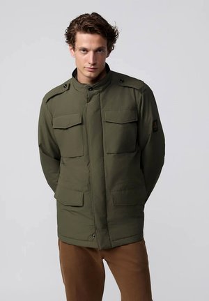Giovane uomo con capelli ricci che indossa una giacca verde in stile militare e pantaloni marroni, in piedi con le mani dietro la schiena su uno sfondo grigio.