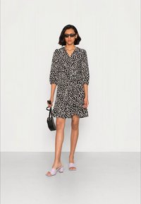 Vestido preto com pontos brancos, decote em V, mangas até o cotovelo, cintura ajustada, comprimento até o joelho, combinado com sandálias de salto bloco lavanda e bolsa preta.