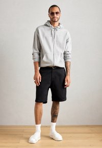 Šedá mikina na zips s šnúrkou, čierne džínsy šortky, biele slip-on topánky a slnečné okuliare. Obsahuje viditeľné tetovania na ruke.