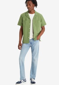 Uomo che indossa una camicia a maniche corte verde chiaro con bottoni sopra una maglietta bianca, jeans azzurri chiari e sneakers blu navy, in piedi con una mano in tasca.